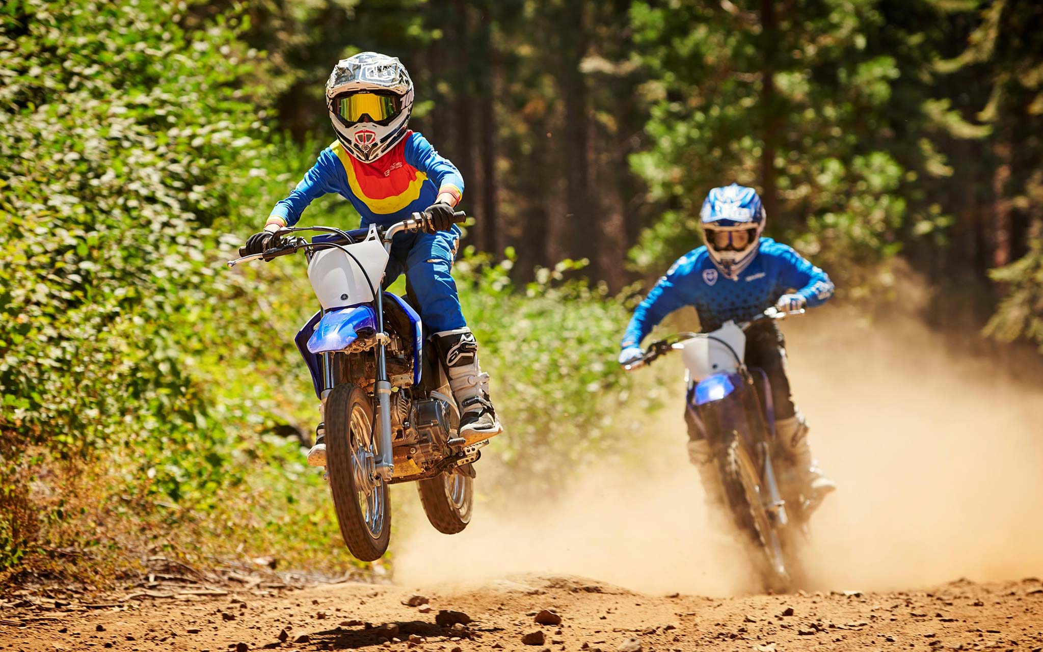 youth on dirt bike taking small jump ahead of adult rider in dust cloud